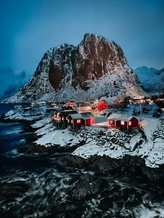 Small village of red cabins next to the sea, with a snowy mountain behind them.